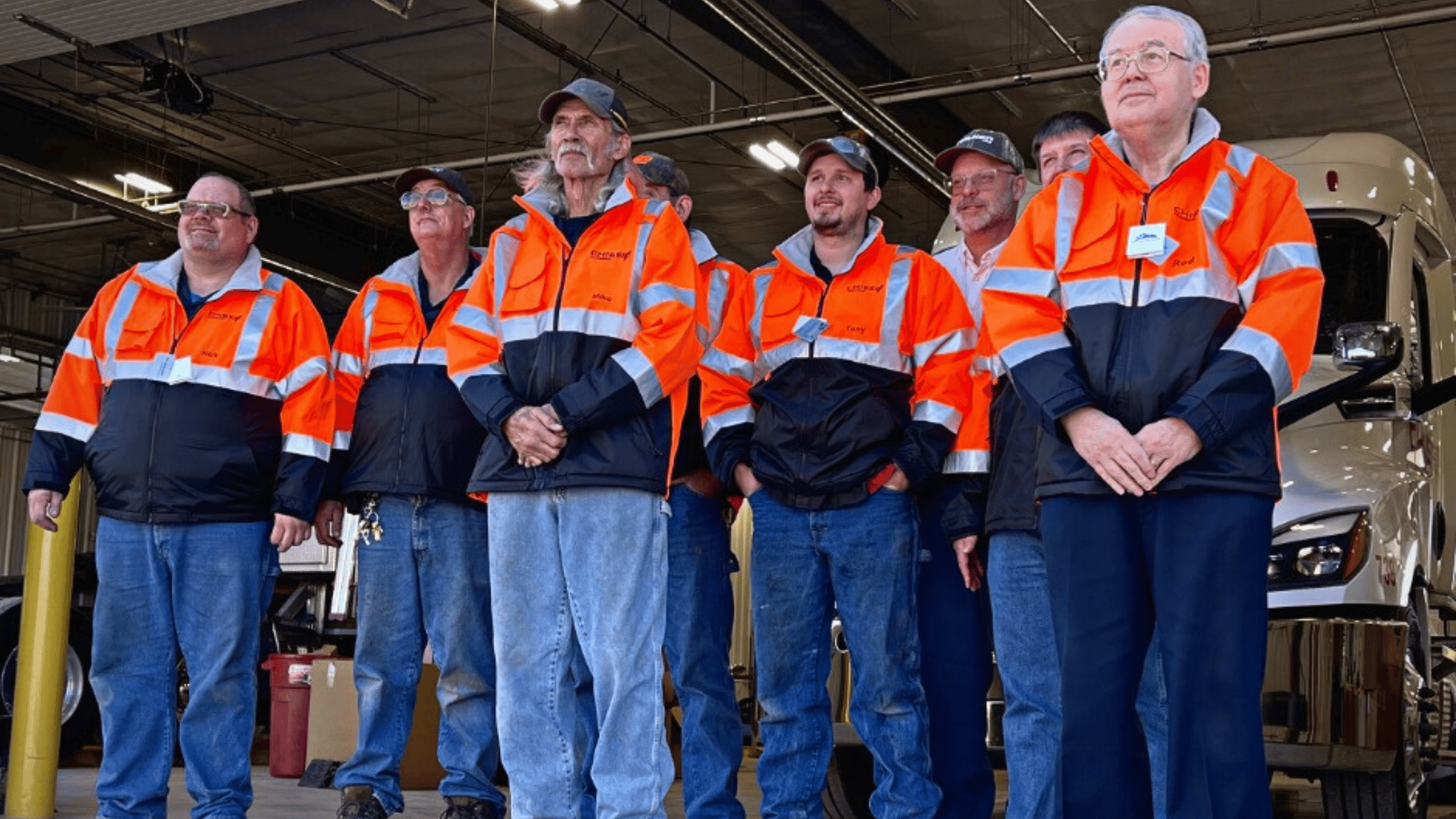 a group of men with blue jeans and orange high vis jackets standing inside of a diesel mechanic shop.