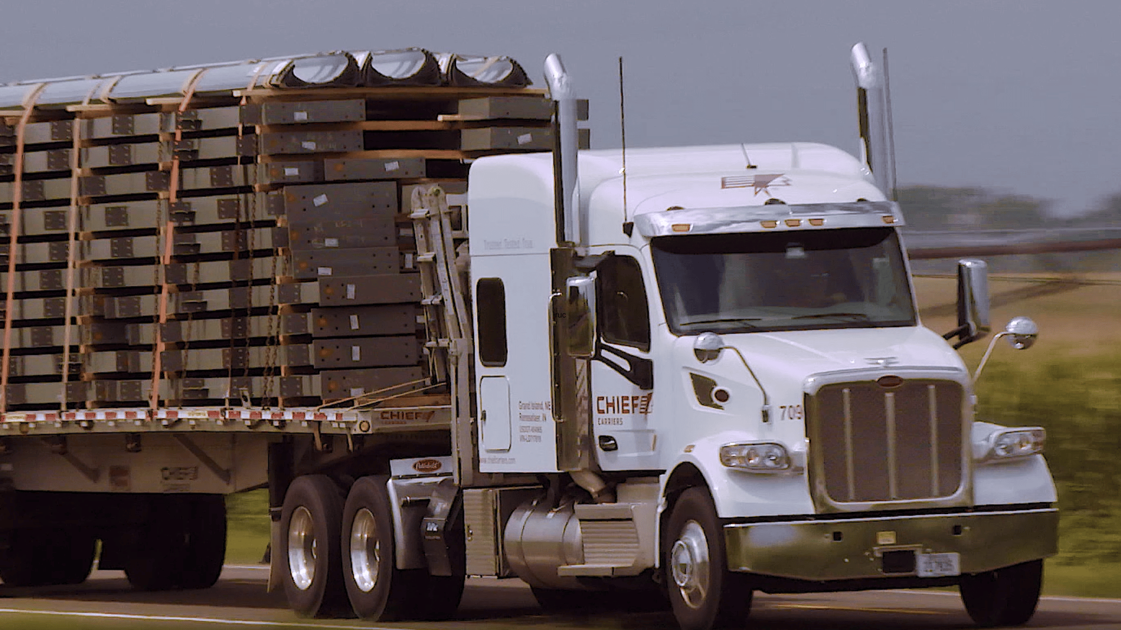 a white flat bed semi truck driving down the highway carrying a load of steel.