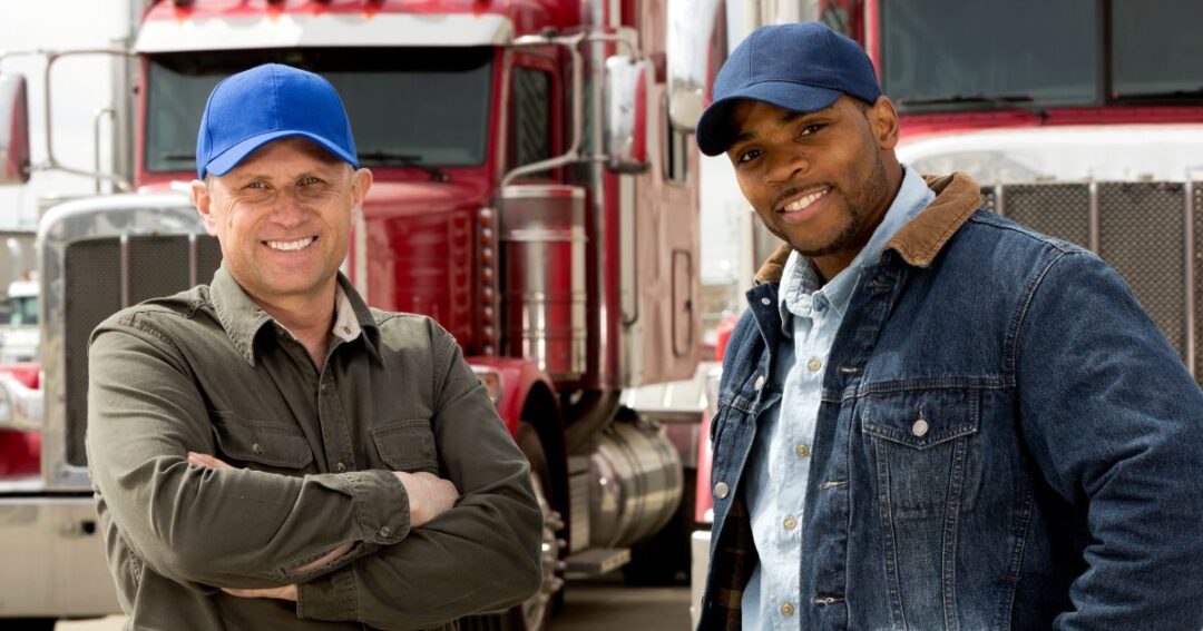 Two smiling truck drivers with red truck cabs behind them.
