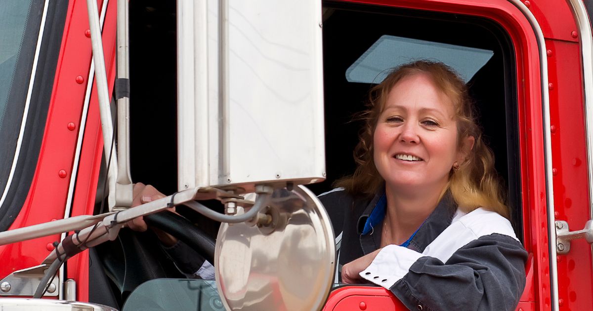 A smiling female driver behind the wheel of a red semi-truck.