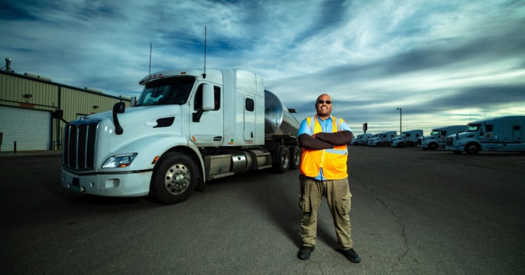 A male truck driver is standing on a lot where a trucking fleet is parked.