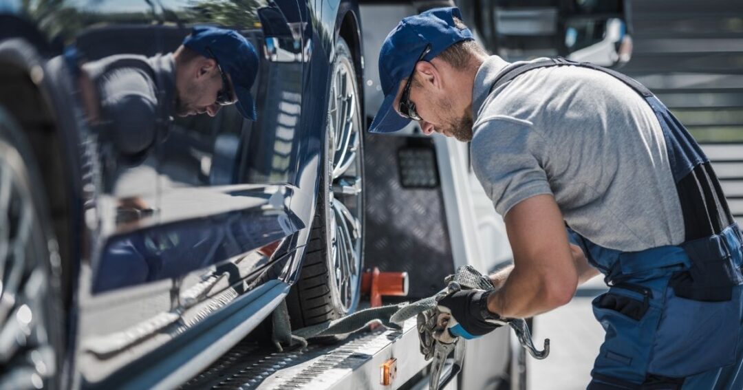 Flatbed trucker driver inspecting strap tie-downs for a car being hauled.