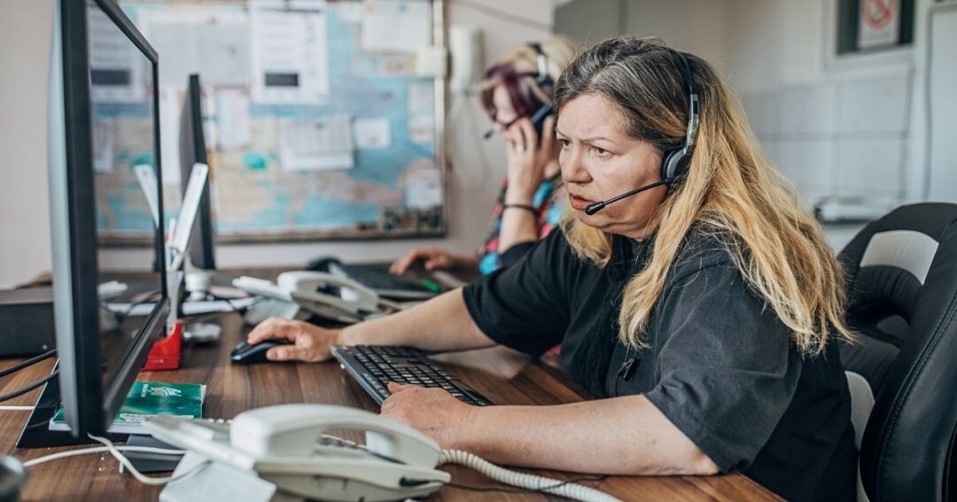 A woman speaking on a headset while getting information from a computer monitor.