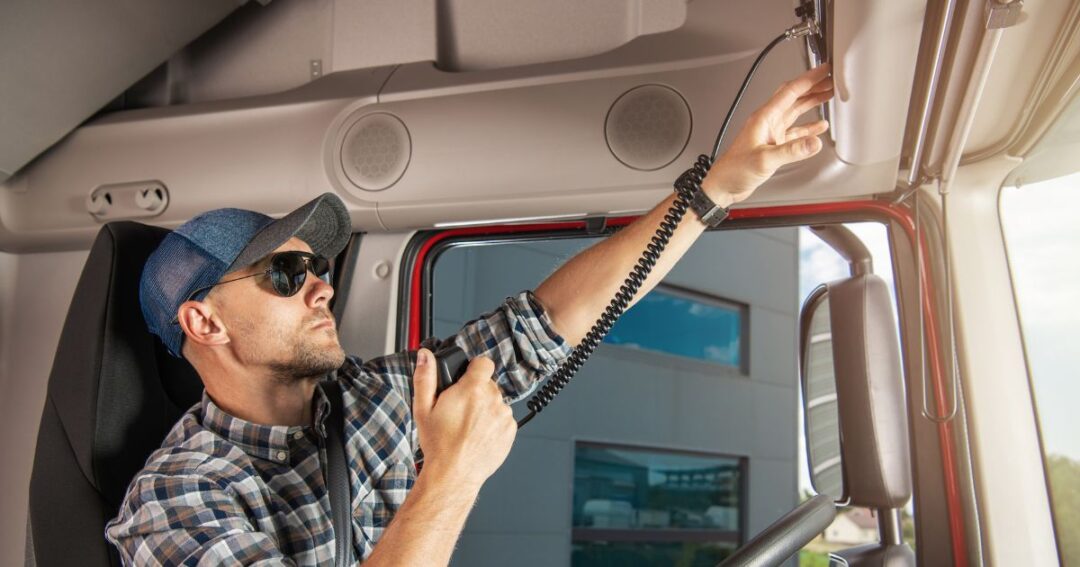 A male truck driver reaching for the radio controls in the truck cab.
