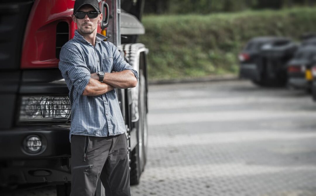 A trucker driver standing by his truck waiting with arms crossed.