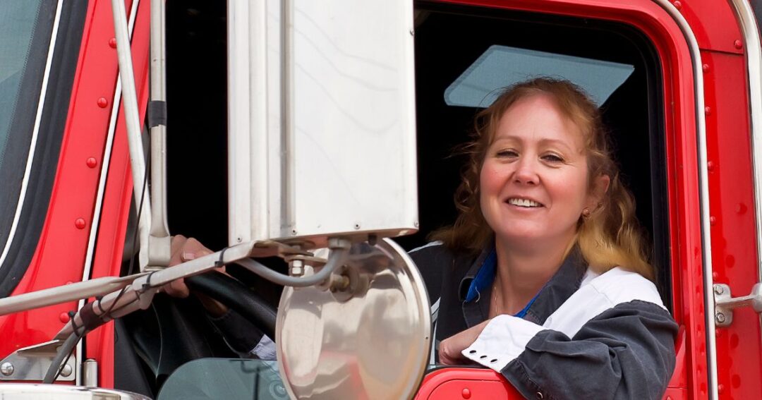A smiling female driver behind the wheel of a red semi-truck.