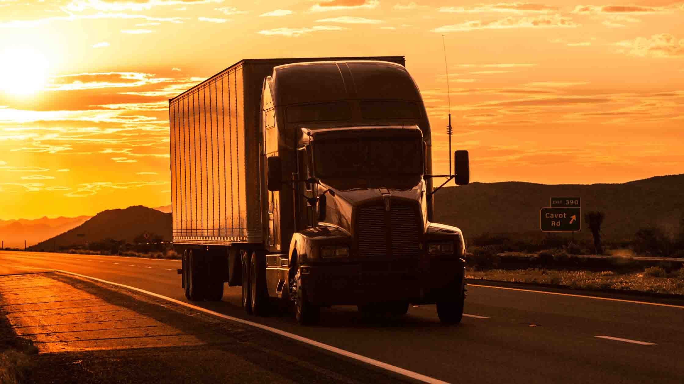 a white semi-truck trailer driving down the highway with the sun setting behind it.