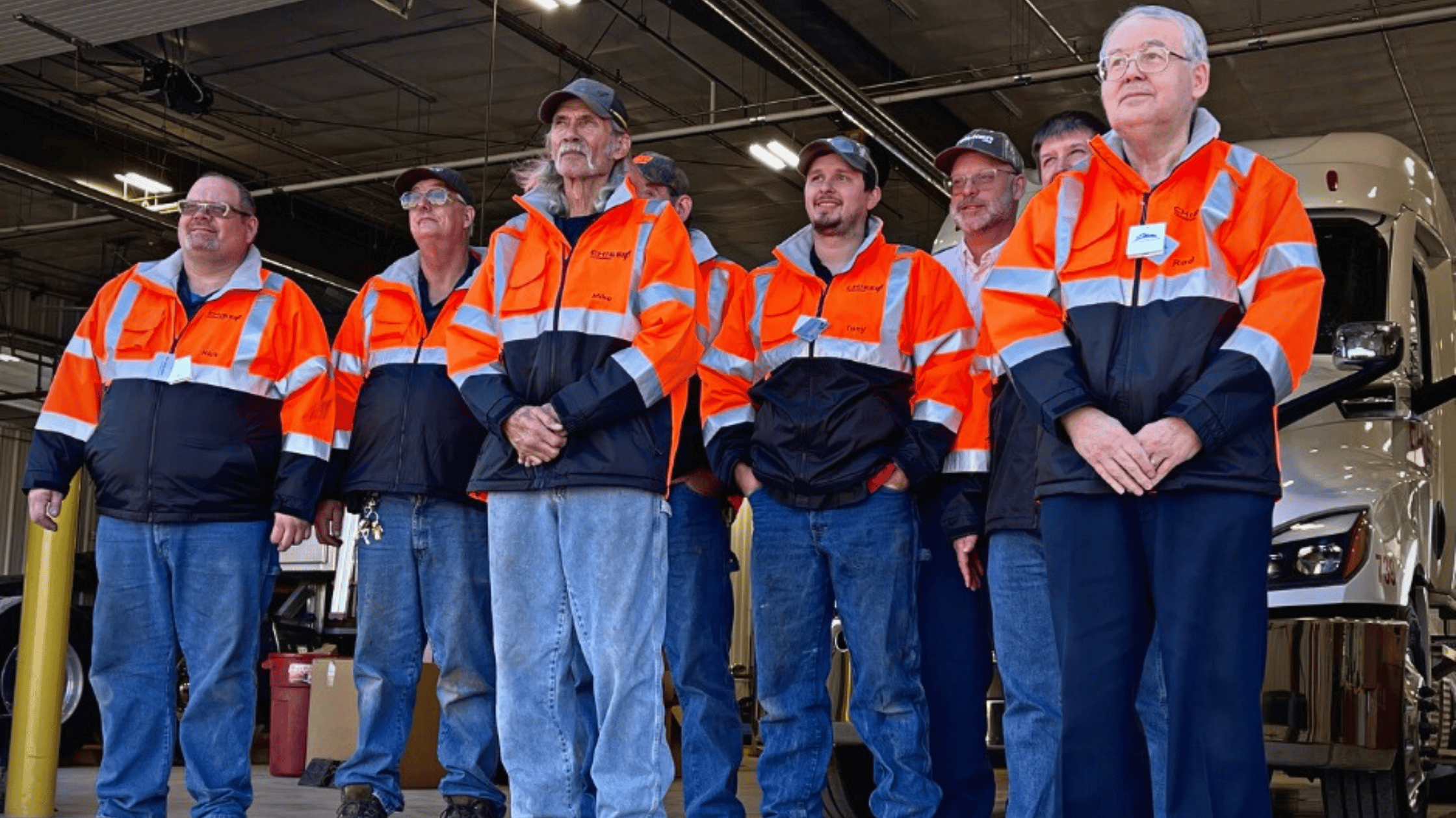 national technician appreciation week a group of men with blue jeans and orange high vis jackets standing inside of a diesel mechanic shop.